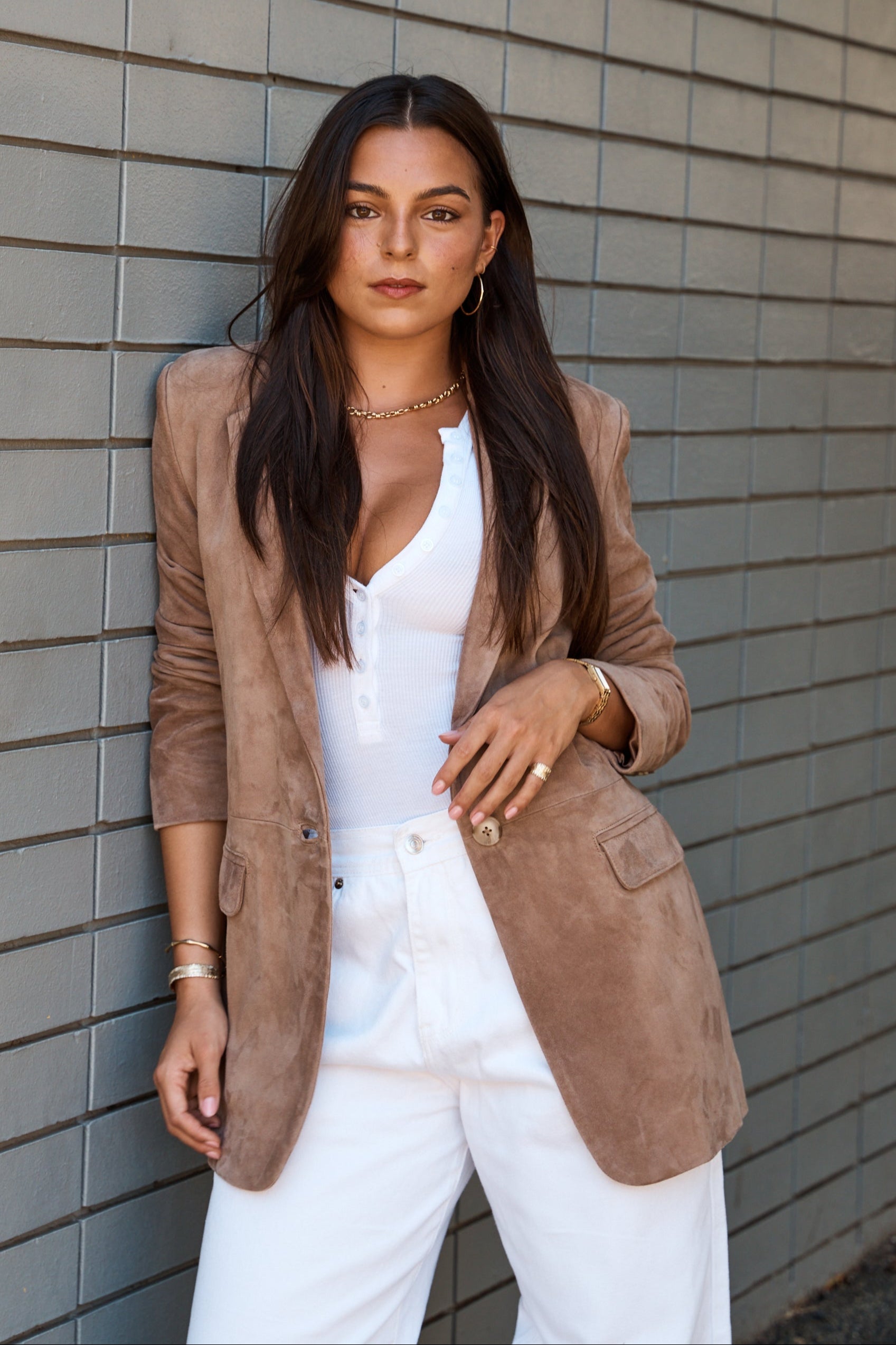 A woman wearing a brown oversized suede blazer with a relaxed fit. She is paired with a white top and white trousers, standing against a gray background.